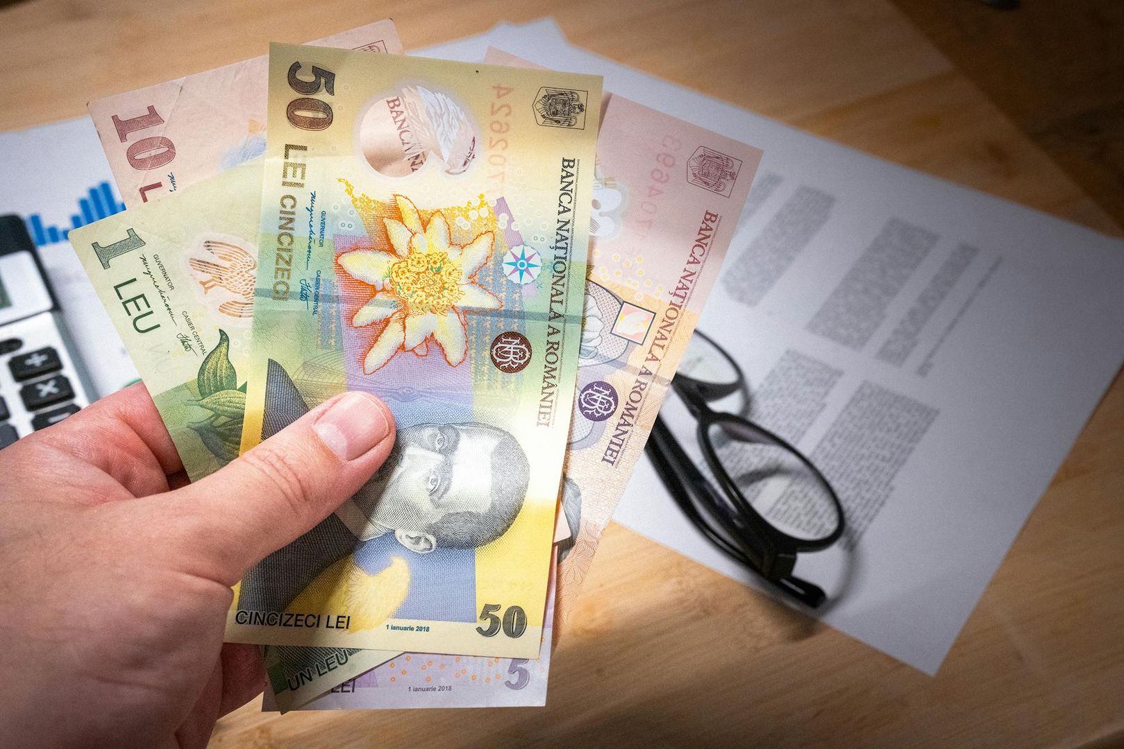 Hånd holder rumænske pengesedler, med regnemaskine og papirer i baggrunden.